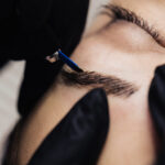 eyebrow microblading. A master in black gloves holds a blending needle over the brow of the model. Macro photography